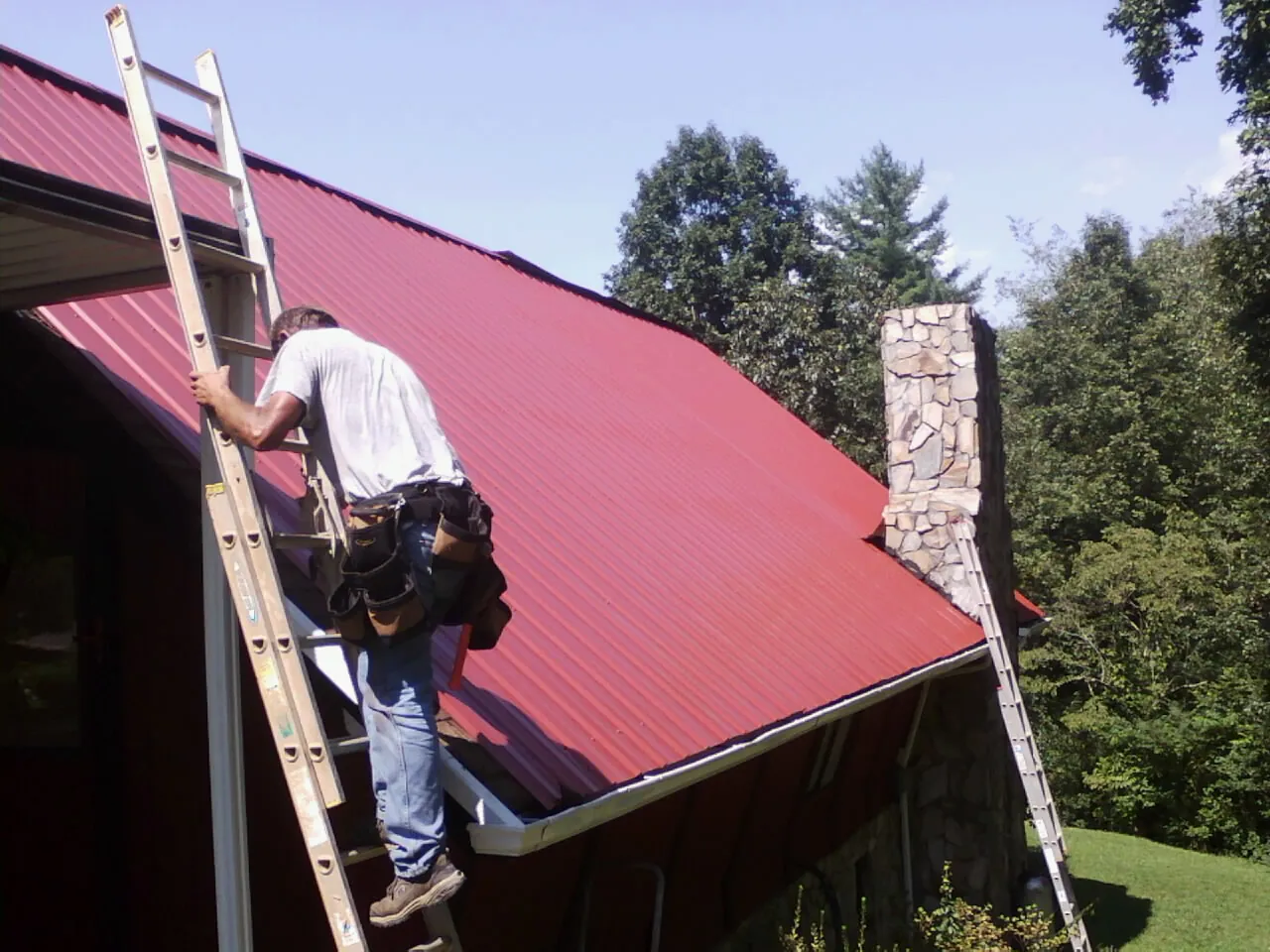 Roofer on a ladder beside a red metal roof and stone chimney — Soffit & Fascia Repair by Roofing Brothers in La Grande